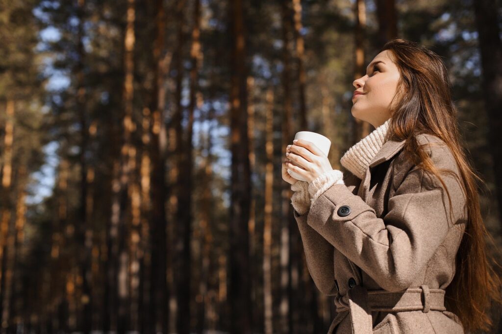 Woman standing in front of pine trees wearing a winter coat and gloves, holding a cup of coffee, symbolizing warmth, relaxation, and enjoying nature Therapy for adults in Blaine MN