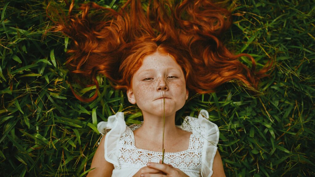 Young redheaded girl lying in the grass with a blade of grass in her mouth, symbolizing playfulness, curiosity, and connection with nature . Therapy for children in Blaine MN