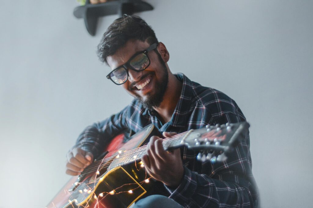 Smiling man joyfully playing an acoustic guitar indoors with fairy lights, symbolizing creativity and joy. Therapy for anxiety in blaine mn