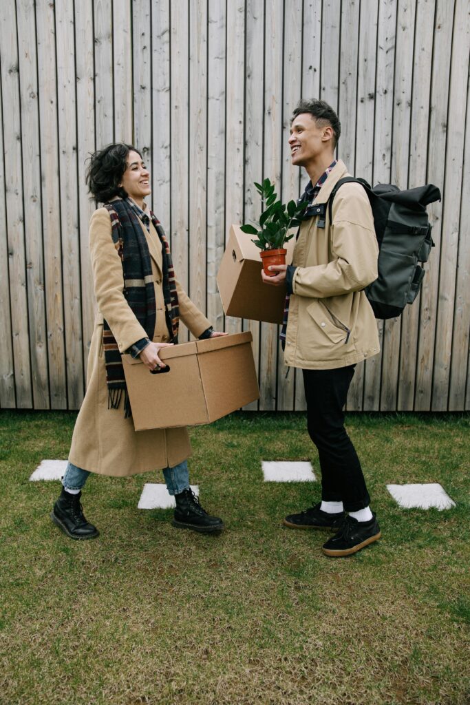 Two people outside carrying moving boxes, one holding a plant, symbolizing life transitions and new beginnings