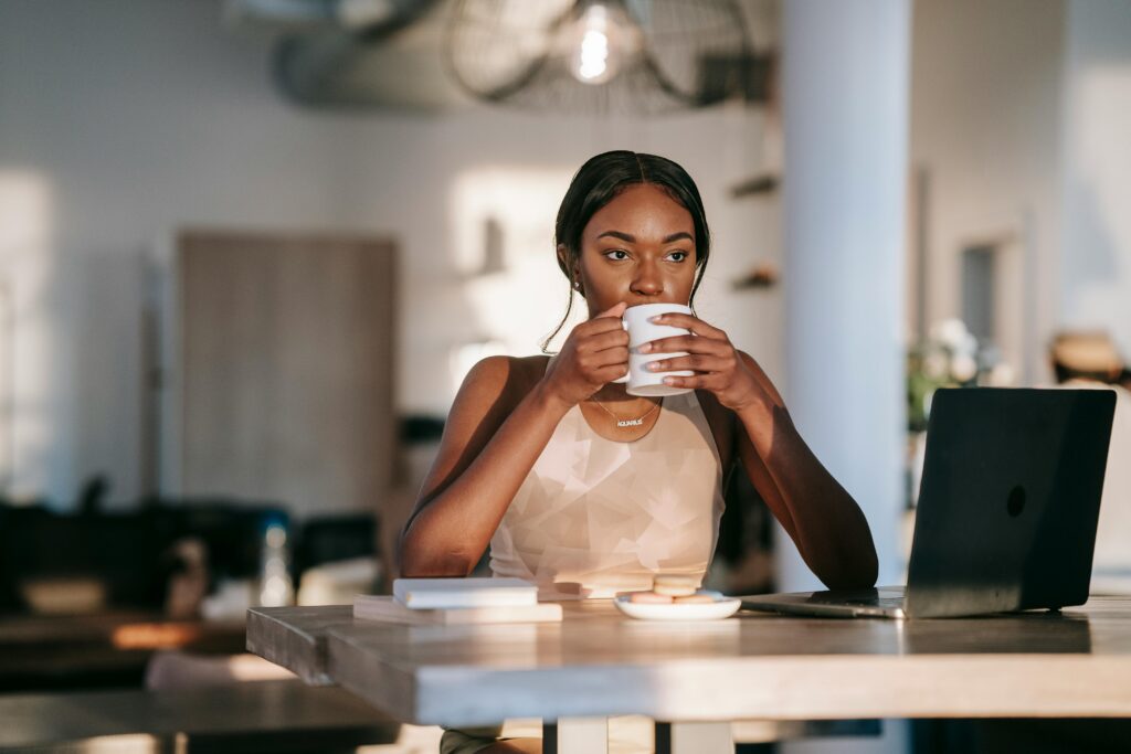 Woman at a table with her laptop open, drinking coffee and looking out into space, symbolizing reflection and calm focus Therapy for adults in Blaine MN