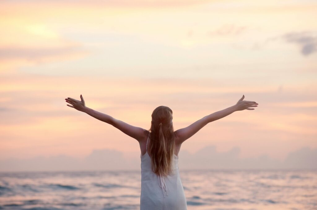 Woman facing the ocean with her arms out wide at sunset under a cloudy pink sky, symbolizing freedom, release, and reflection. Therapy For Life Transitions In Blaine MN