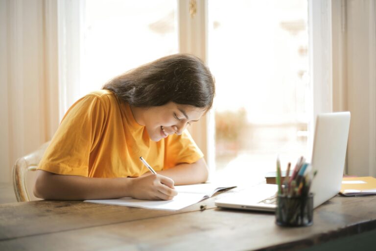 Woman smiling while sitting in front of a bright window, writing in a notebook with her laptop open, symbolizing productivity and positivity Therapy For Anxiety in Blaine MN