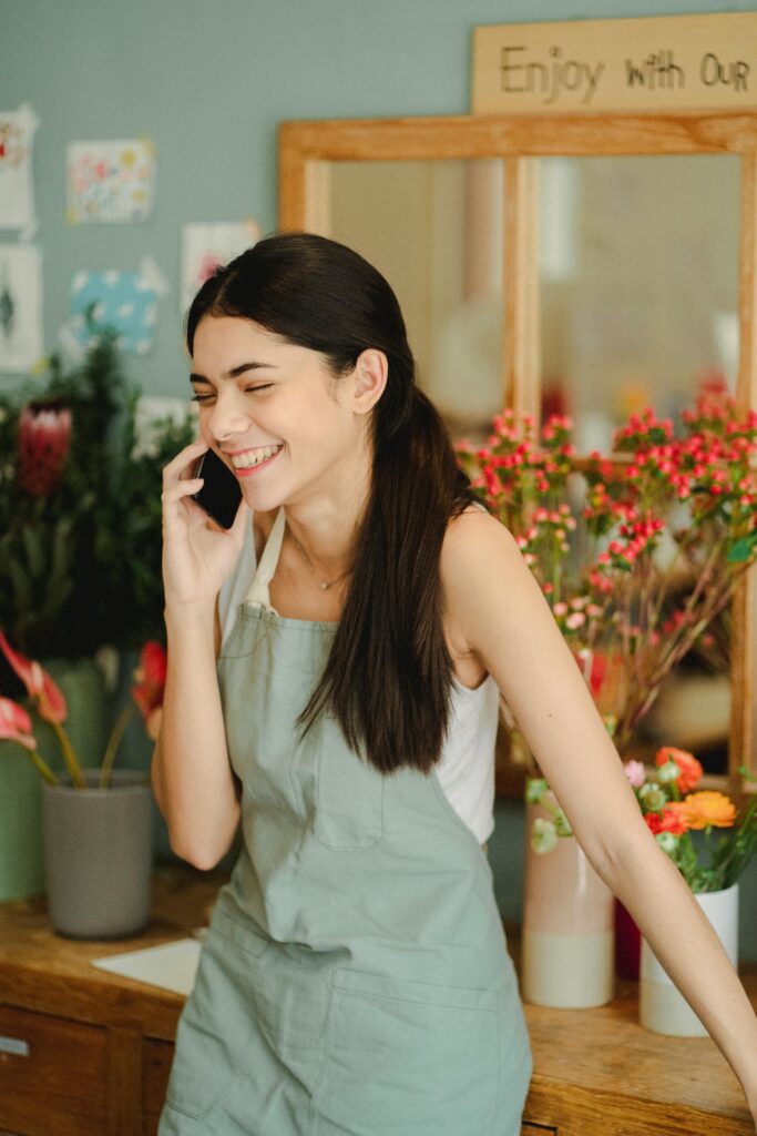 Cheerful female worker with closed eyes wearing apron standing near wooden table with flowers in vase while talking on smartphone in floristry studio. Therapy for self esteem in Blaine MN