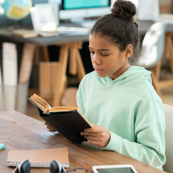 Cute elementary schoolgirl in casualwear sitting by table in living-room against workplace with computer and reading school literature