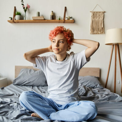 appealing queer person with vibrant red hair sitting on bed and looking away, leisure time