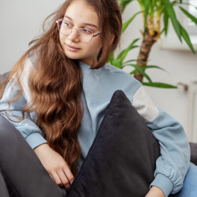 Thoughtful teenage girl wearing glasses relaxing on a sofa, looking out the window with a calm, pensive expression.