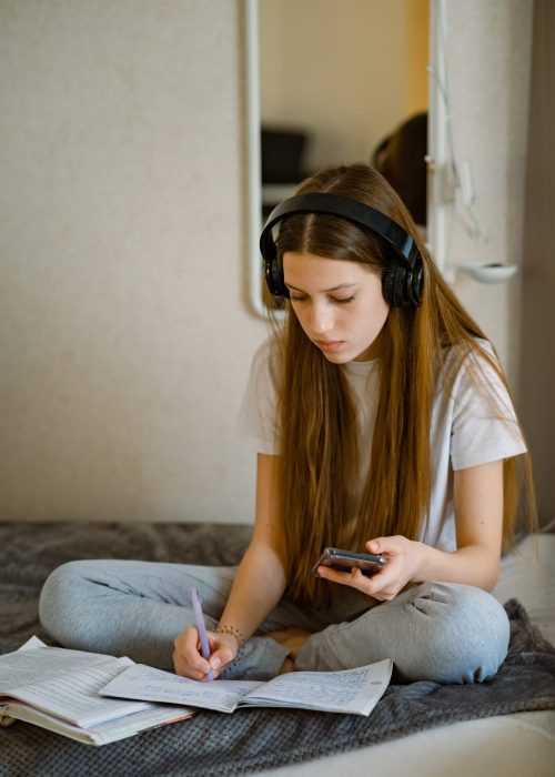 Teenage girl at home on the bed in headphones and with a smartphone, lined with textbooks and terady, to study, prepares lessons.