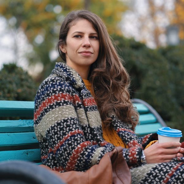 Pretty young woman in good mood enjoying coffee in the autumn day, while sitting on a bench, enjoying the good weather.