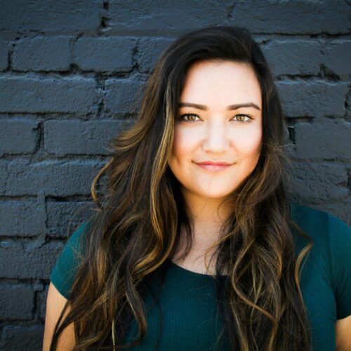 Woman with brown hair smiling at the camera, standing against a black wall wearing a black t-shirt, symbolizing confidence and approachability Therapy for adults in Blaine MN