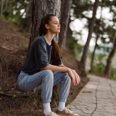 Serene young woman dressed in comfortable attire, sitting against a tree along a stone pathway Surrounded by greenery, she embodies tranquility and relaxation in nature