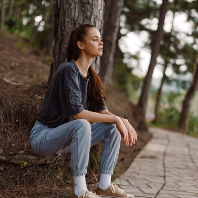 Serene young woman dressed in comfortable attire, sitting against a tree along a stone pathway Surrounded by greenery, she embodies tranquility and relaxation in nature