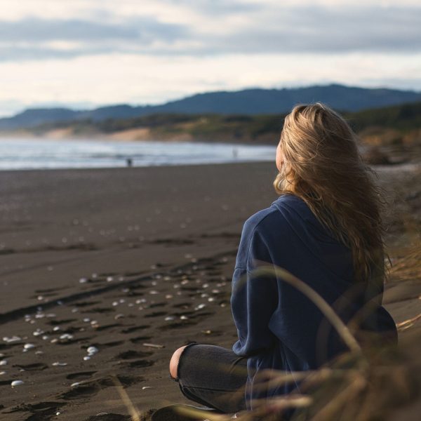 A young woman sits on the shore at sunset, gazing out into the glimmering sea